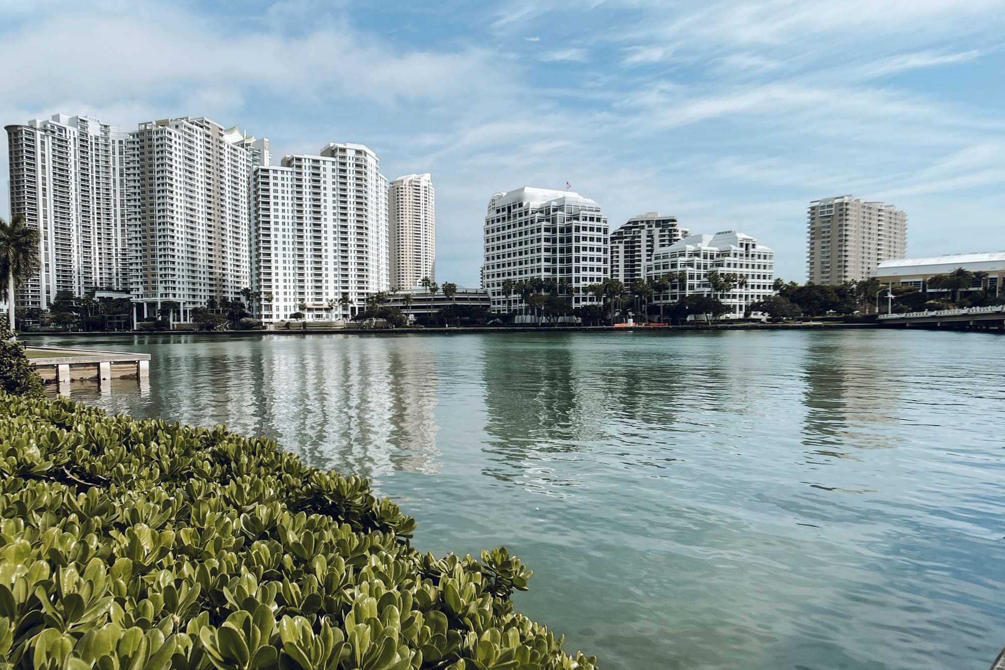 A row of white residential high-rise buildings reflected in calm water under a blue sky, in South Florida